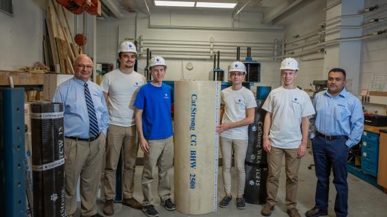 Group of Civil Engineering students wearing white hard hats with blue paw prints on them, flanked by two faculty members, holding a roll of material printed with "CatStrong CG BHW 2500."
